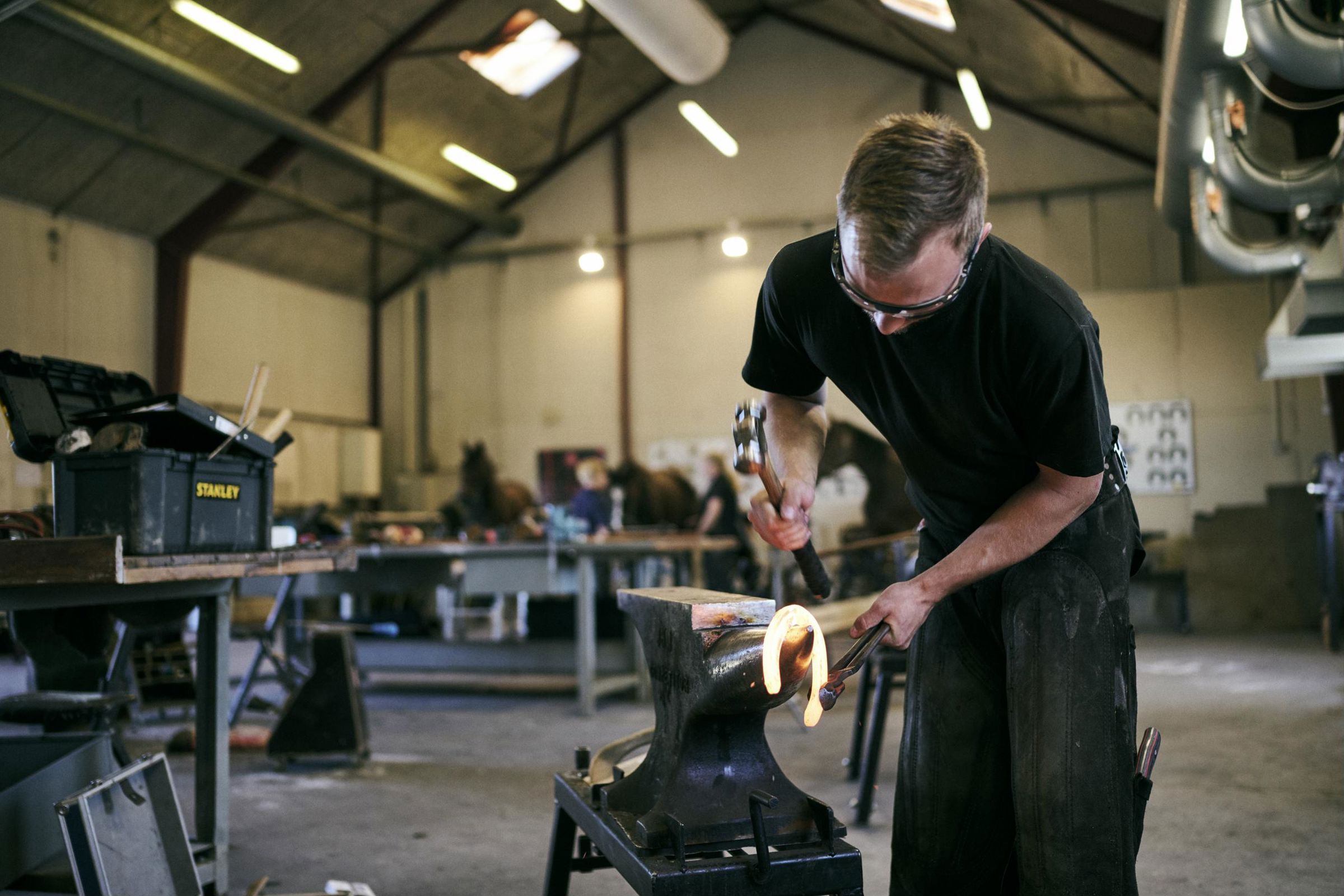 En beslagsmedelærling er ved at smede en gloende varm hestesko i Hansenbergs beslagsmedje.
