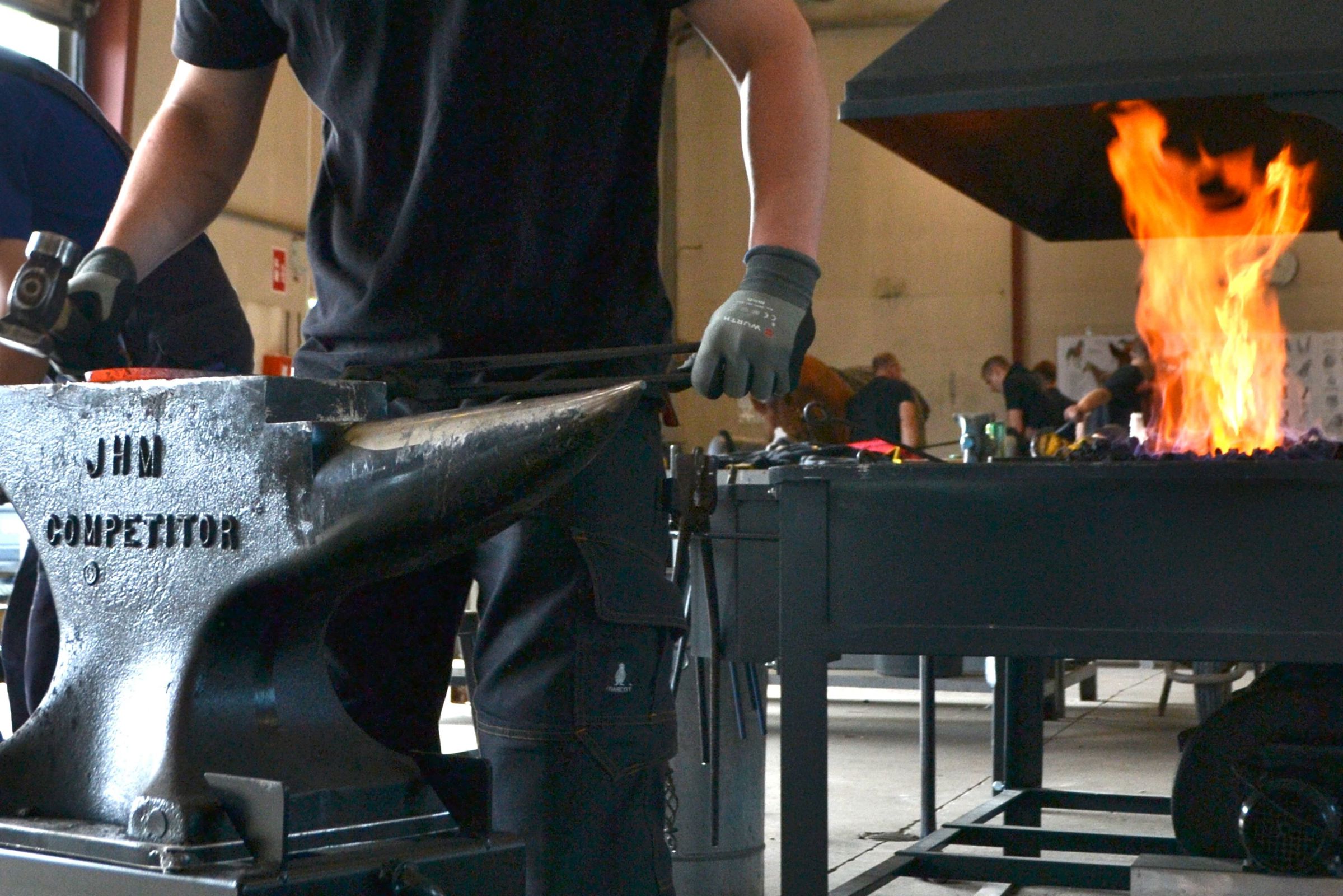 En beslagsmedeelev bruger ambolten, når de håndsmeder en hestesko, og nogle gange bruger de også den store esse i baggrunden, når de varmer hesteskoen op i Hansenbergs beslagsmedje.